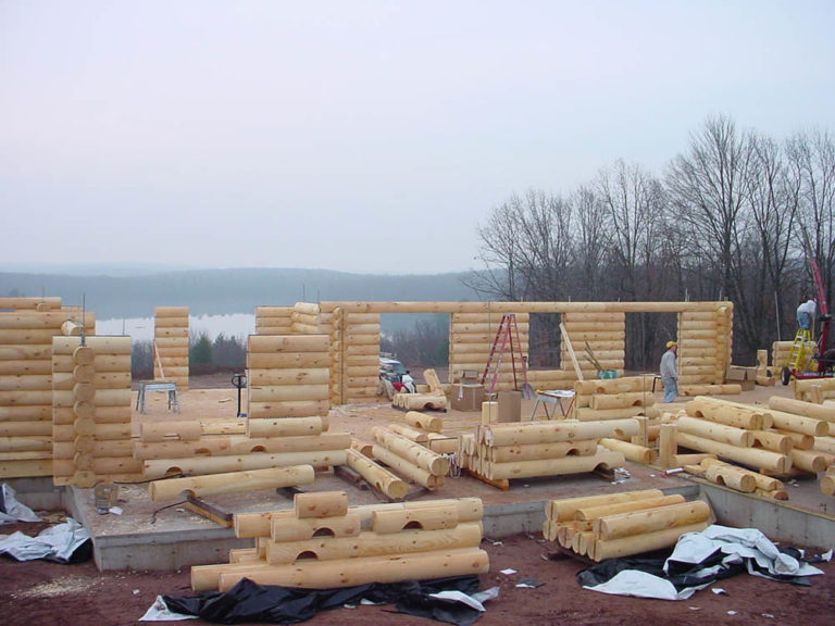 Log home construction site featuring stacked logs, building framework, and workers, showcasing Estemerwalt Lumber Products' commitment to quality craftsmanship and lumber delivery services.