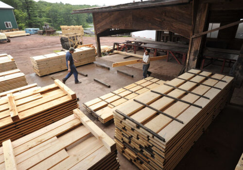 Workers handling lumber in a rustic lumber yard, showcasing stacks of Eastern White Pine boards and timbers available for various construction and decorative uses.