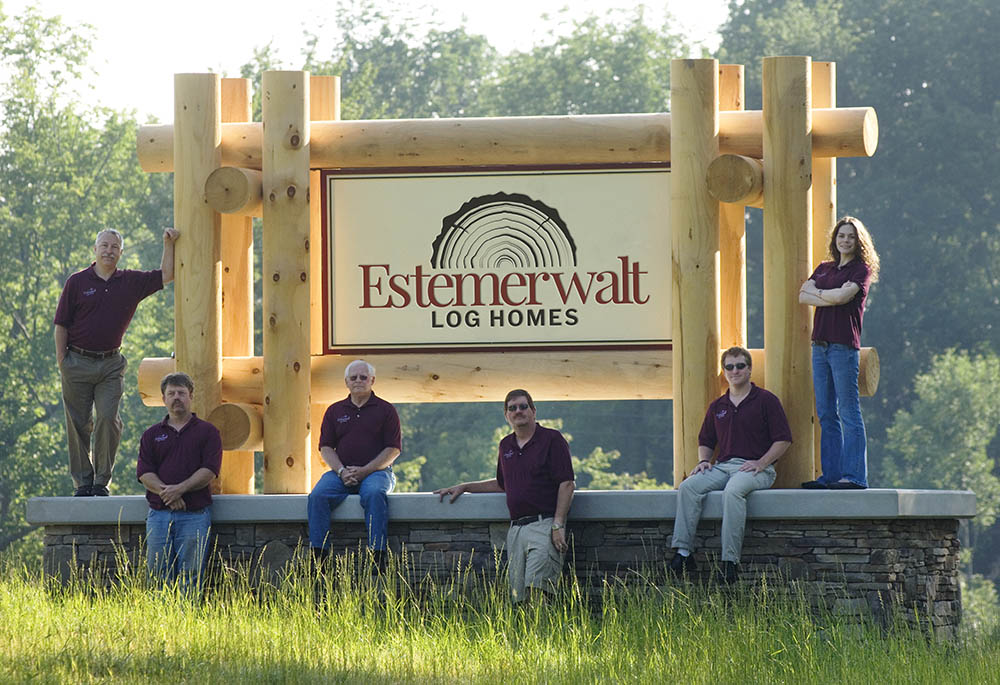 Estemerwalt Log Homes team members posing in front of a prominent wooden sign displaying the company name, surrounded by greenery, emphasizing community and craftsmanship in log home manufacturing.